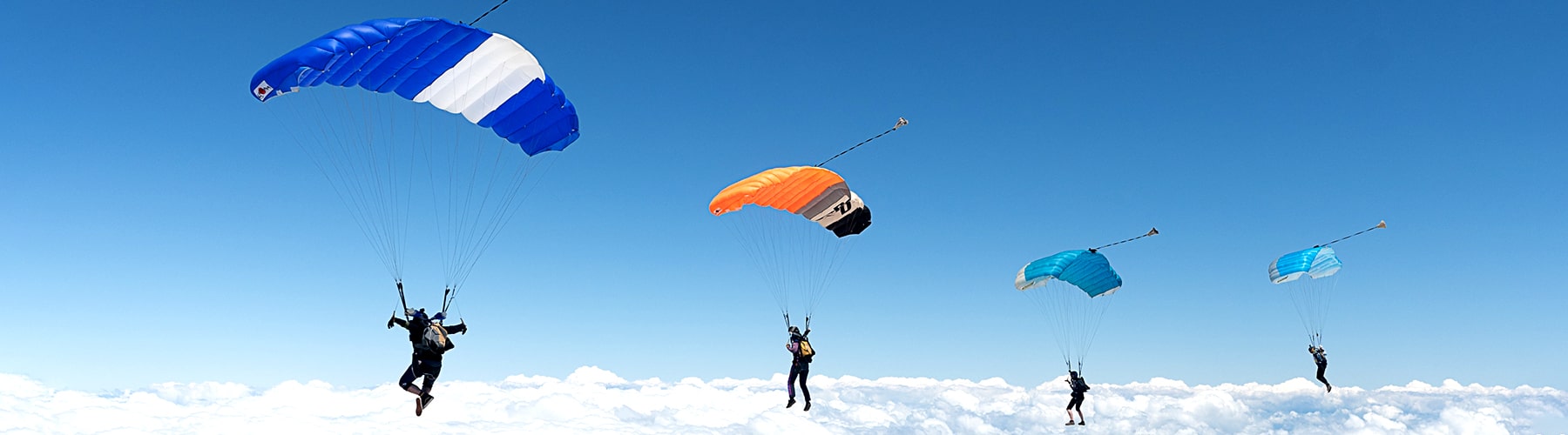 A group of four skydivers flying with colourful canopies above the clouds.