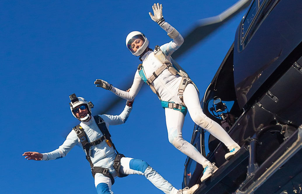 A man and a woman in white flight suits jumping out of a black helicopter.