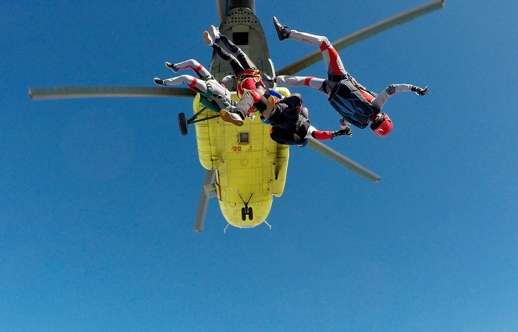 Three skydivers in freefall after jumping out of a yellow helicopter.