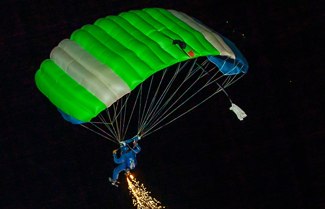 Person skydiving at night with a green parachute, trailing pyrotechnics.