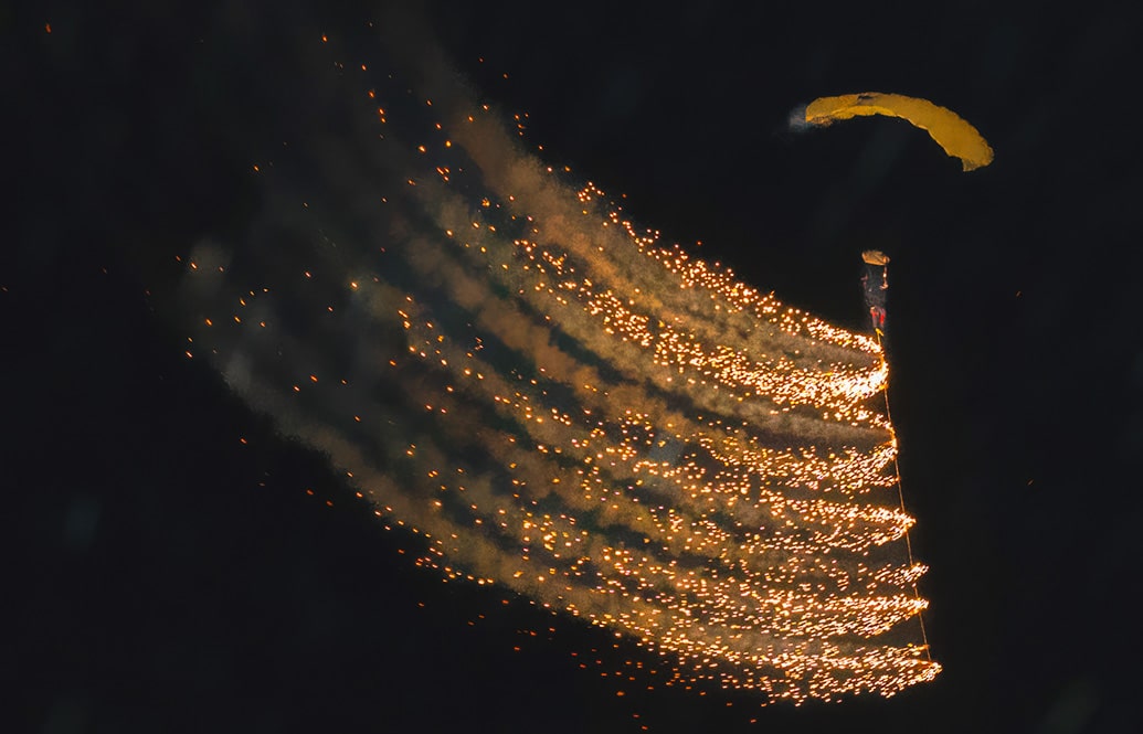 Spectacular fireworks being trailed behind a skydiver under canopy in a dark sky.