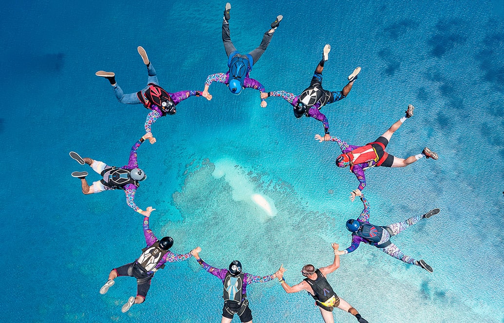 A ring of skydivers in freefall above blue water.