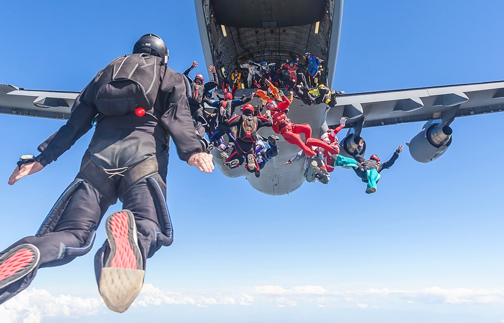 A load of colourful skydivers jumping out of the rear cargo door of a military aircraft.